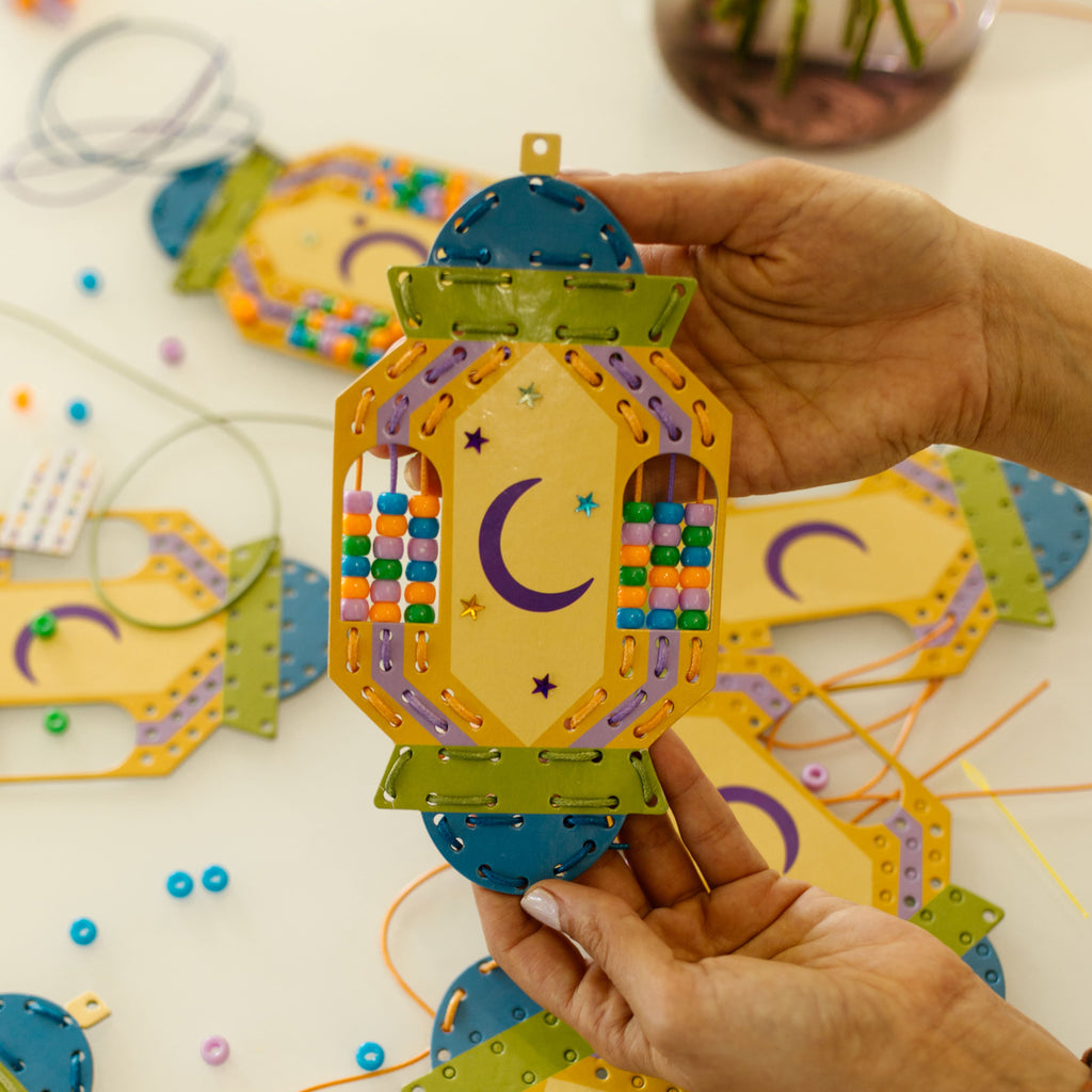 Hand holding a colorful paper eid lantern craft with more ramadan lanterns on a white surface.