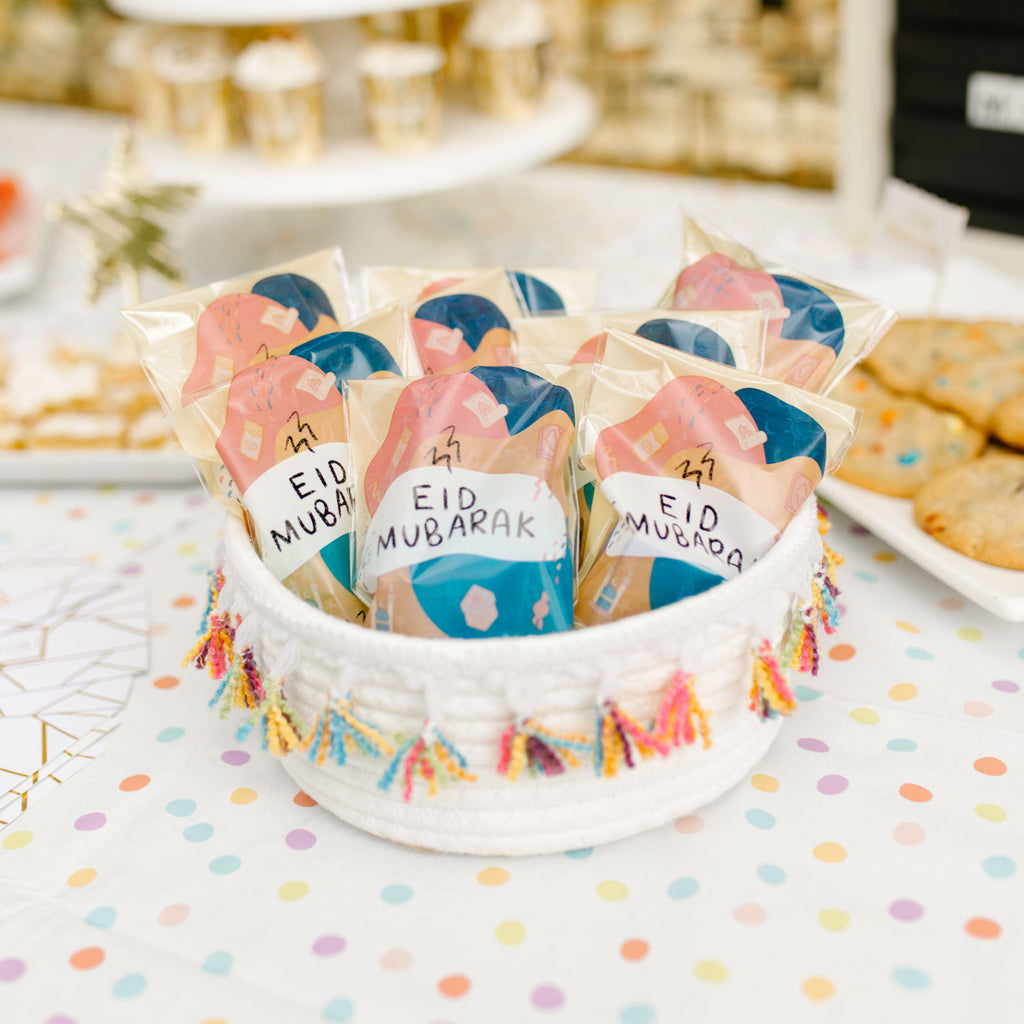 Basket of colorful 'Eid Mubarak' bags on a polka dot tablecloth with blurred dessert display in the background.