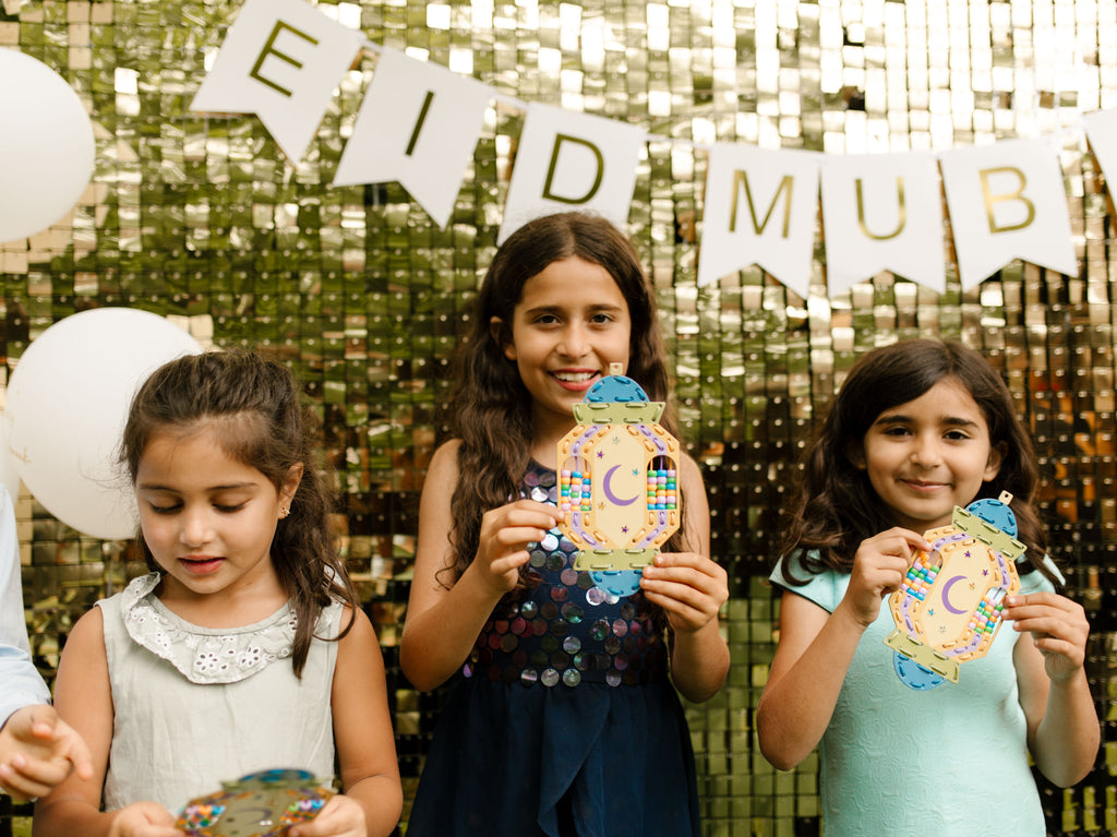Three children holding decorated craft in front of an 'EID MUBARAK' banner with a glittery gold background.