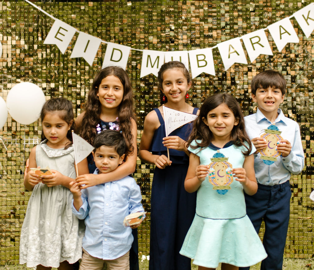 Children standing in front of 'Eid Mubarak' banner with balloons outdoors.